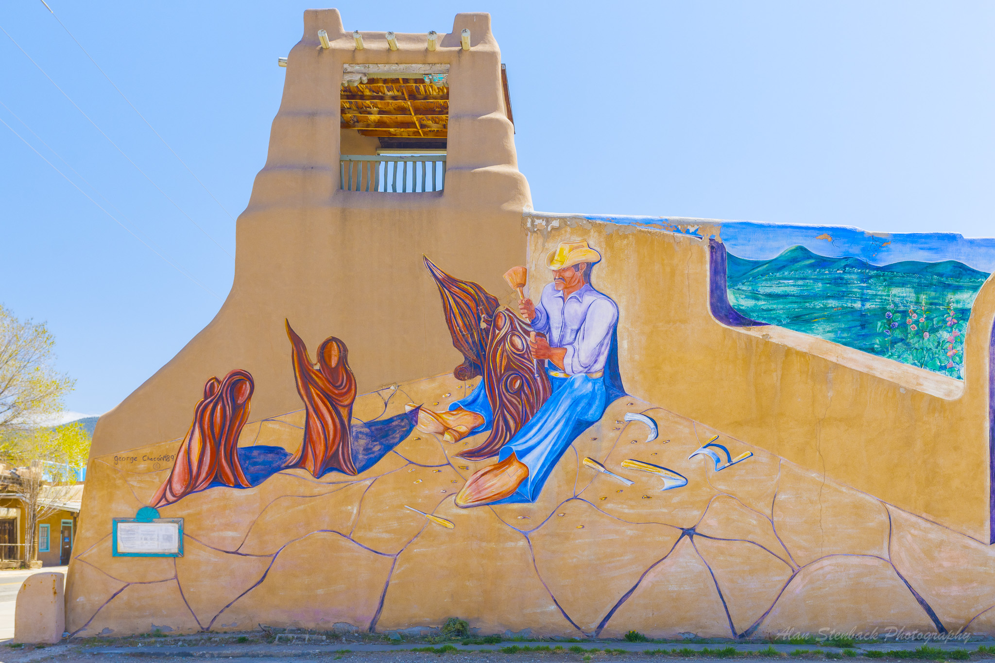 Colorful mural on an adobe building depicting a man in a hat carving figures, surrounded by vibrant landscapes and abstract shapes.