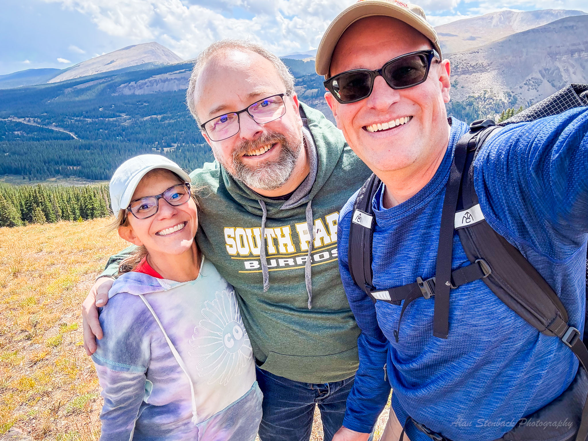 Three friends smiling on a scenic mountain hike with a forested landscape and blue skies in the background.