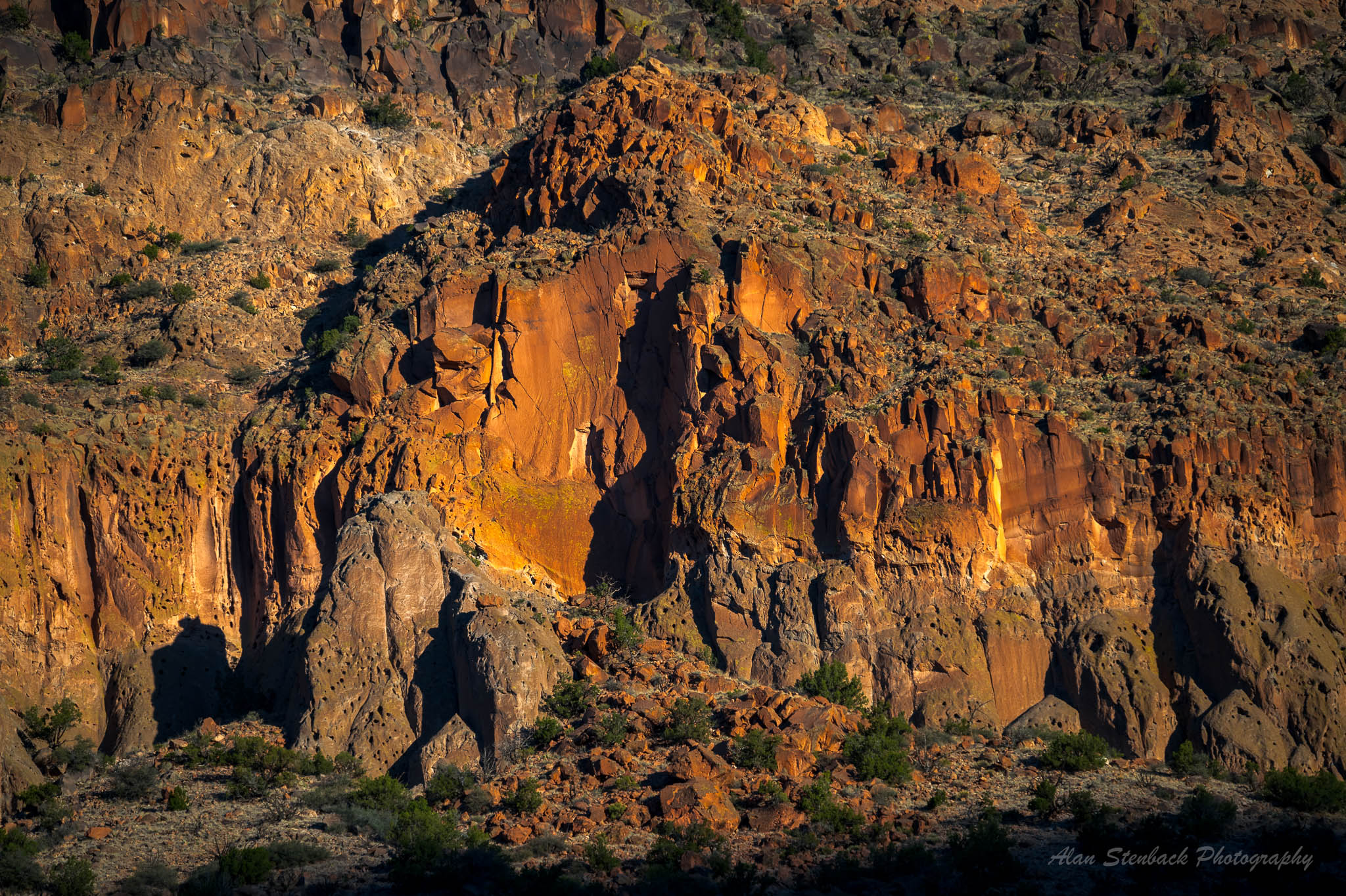 Sunlit orange and brown cliff face with rocky texture and scattered vegetation, showcasing natural geological formations.