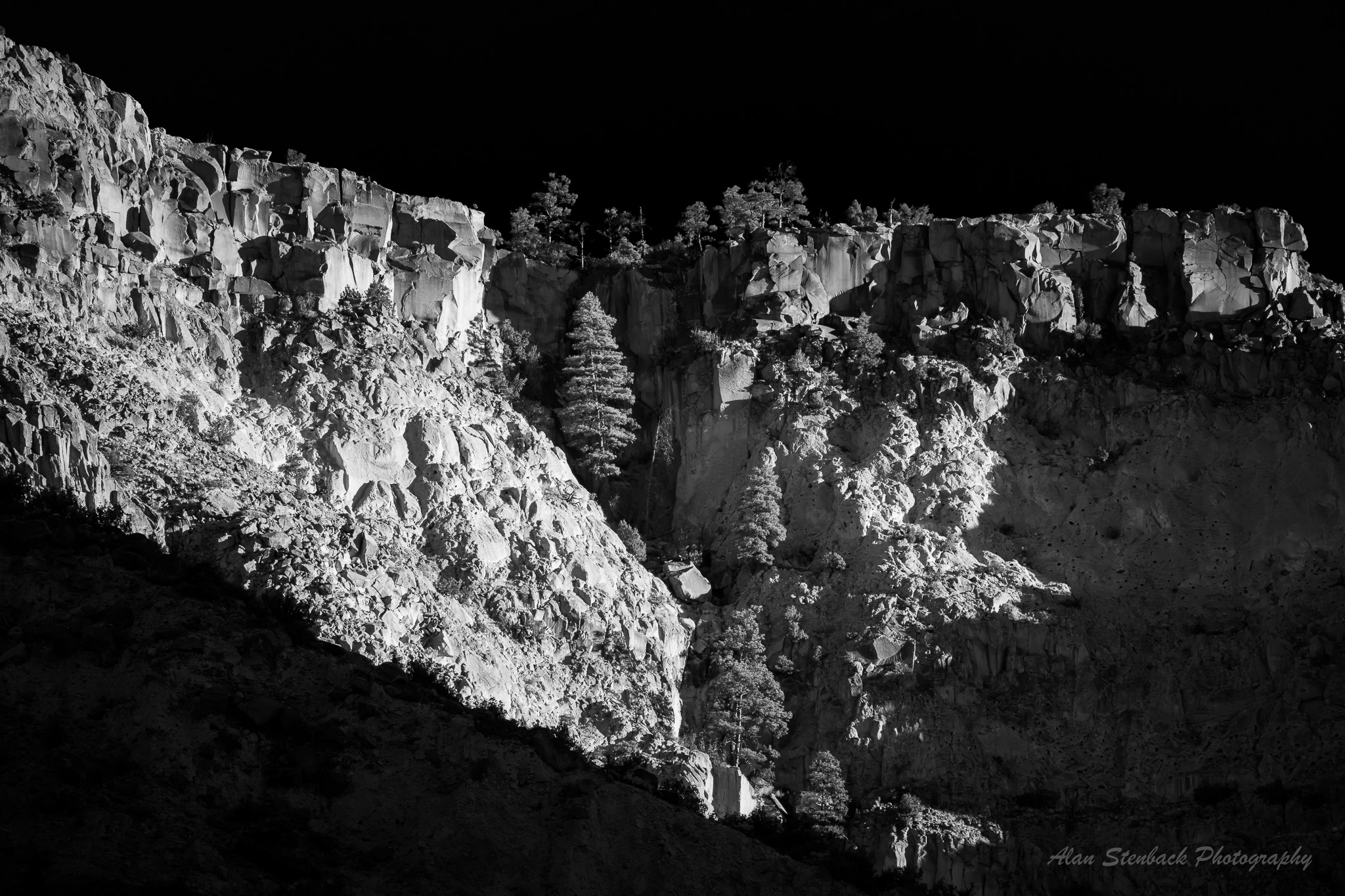 Dramatic black and white cliff landscape with stark contrasts, rugged terrain, and scattered trees under a dark sky.