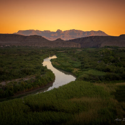 Sunset over winding river surrounded by lush greenery, distant mountains silhouetted against a vibrant orange sky.