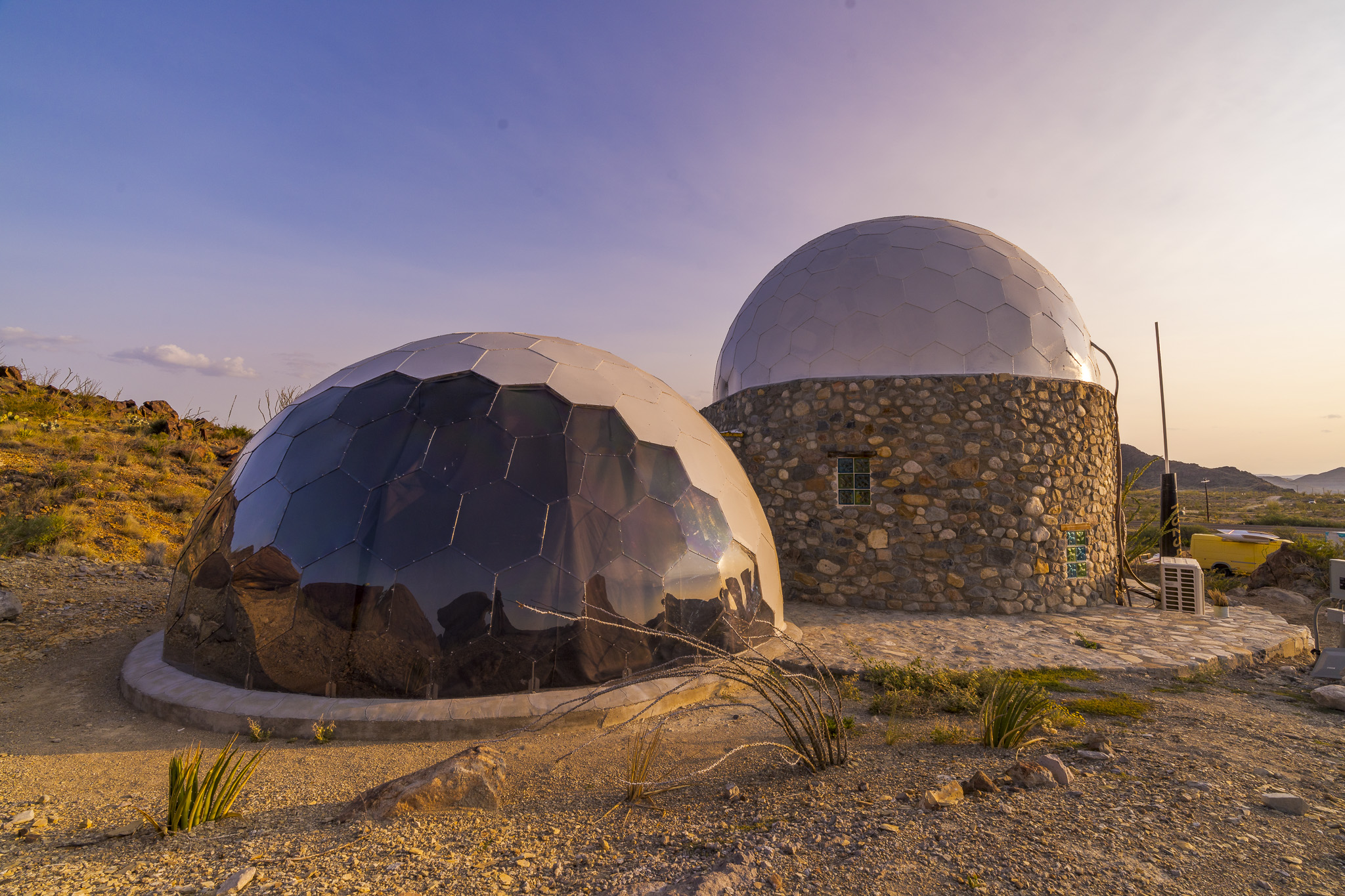 Geodesic domes in desert landscape at sunset, featuring a reflective dome and a stone structure with smooth, rounded shapes, surrounded by rocky terrain and sparse vegetation.