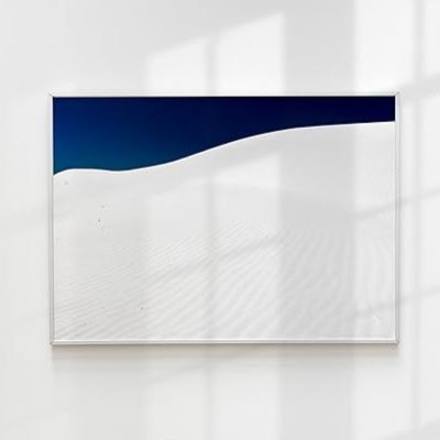Framed desert landscape photograph with white sand dunes and deep blue sky, displayed on a white wall with window shadows.